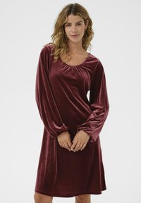 Naised laineliste blondide juustega, kes kannab pikka burgundi värvi sametkleiti, seisab käed õrnalt koos ees ühtlasel taustal.