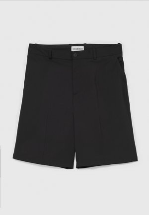 Musta lyhythihainen nahkapaita etutaskulla, yhdistetty mustiin shortseihin, mustiin polvisukkiin ja tummiin slip-on-kenkiin. Yksinkertainen, moderni muotoilu.