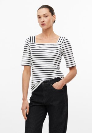 Femme portant un haut blanc à rayures noires avec un col carré et des manches aux coudes, et un pantalon noir taille haute, main dans la poche.
