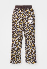 Leoparda raksta sporta bikses brūnā un dzeltenā krāsā ar auklu jostu un baltu "alpha industries" tekstu kreisajā kājā.