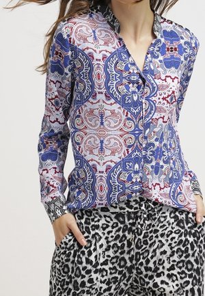 Frau trägt eine langärmelige Bluse mit blau-rotem und weißem Paisley-Muster und schwarze und weiße Leoparden-Print-Hose, Hände in den Taschen.