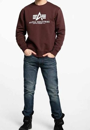 Mann trägt ein braunes Alpha Industries Sweatshirt, blaue Jeans und schwarze Stiefel, steht mit den Händen in den Taschen vor einem weißen Hintergrund.