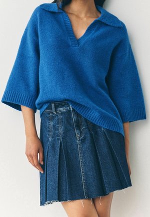 Femme portant un pull en maille bleu ample avec un col en V et une mini-jupe en jean plissée avec un ourlet effiloché.