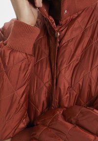 Kurtka pikowana w głębokim rudobrązowym kolorze, z prążkowanymi mankietami i wysokim kołnierzem zapinanym na zatrzaski. Gładki, błyszczący materiał.