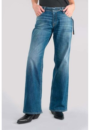 Jean en denim bleu avec une coupe décontractée et des jambes évasées. Présente un design à cinq poches, des passants de ceinture et un léger dégradé. Porté avec des bottines noires.