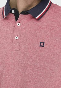 Rode polo met een gestructureerde weving, voorzien van een marineblauwe kraag met rode en witte strepen en een klein marineblauw logo op de borst.