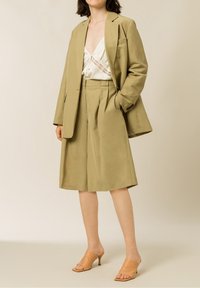 Blazer beige su misura, pantaloni larghi coordinati e blusa crema con dettagli in pizzo; abbinati a mule aperti in tinta sottile. Texture del tessuto liscia.