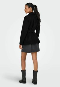 Manteau noir ceinturé avec un design ajusté, présentant un col cranté et une forme évasée, porté sur une mini-jupe grise et des bottines noires.
