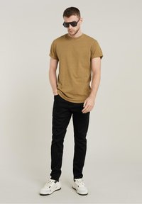 Camiseta de manga corta color caqui, jeans negros y zapatillas blancas. El modelo lleva gafas de sol rectangulares negras. Textura suave, ajuste casual.