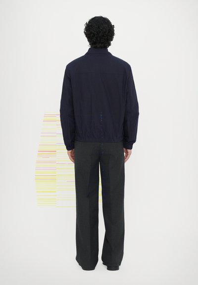 Giacca leggera navy con colletto alto e orlo elastico, abbinata a pantaloni larghi scuri. Texture liscia e design minimalista.