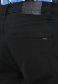 Czarne dżinsy o gładkiej fakturze, z prostokątną skórzaną metką z wytłoczonym napisem "TOMMY JEANS" oraz małym logo w kolorach czerwonym, białym i niebieskim w pobliżu kieszeni.