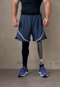 Idrottare bär svart åtsittande tröja, mörkgrå shorts med meshdetaljer och lila sneakers, och visar upp en metallprotes på höger ben.
