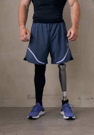 Sportists valkā melnu pieguļošu kreklu, tumši pelēkus šortus ar sieta akcentiem un violetas sporta kurpes, demonstrējot metāla protēzi uz labās kājas.