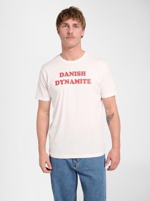 Homme aux cheveux courts et à la moustache, portant un t-shirt blanc avec le texte rouge "DANISH DYNAMITE" et un jean bleu, debout devant un fond uni.