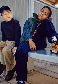 To barn sitter på en benk. Den ene har på seg en marineblå genser og beige bukser. Den andre har en grønn vest over en denimskjorte, blå bukser og sneakers.