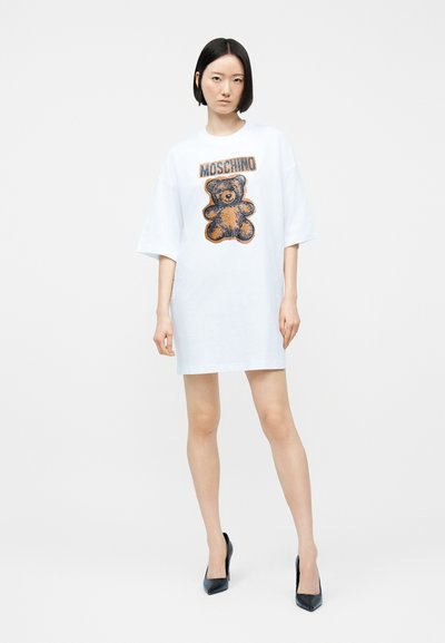 Γυναίκα που φοράει λευκό oversized φόρεμα t-shirt με σχέδιο αρκουδάκι και το κείμενο "MOSCHINO", συνδυασμένο με μαύρα ψηλοτάκουνα, στέκεται μπροστά σε λευκό φόντο.