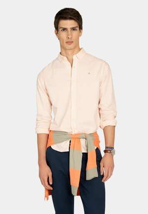 Joven que lleva una camisa de manga larga color durazno claro abotonada, pantalones azul marino y un suéter de rayas enrollado y atado alrededor de su cintura.