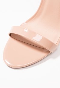 Nude patentti avonaisen varpaan korkokengät, joissa on kiiltävä vyö jalkaterän ylitse ja sileä pinta pohjalla, muotoiltu siluetti.