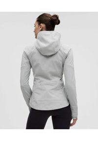 Chaqueta con capucha gris claro de textura suave, diseño ajustado y costuras. Cuenta con un bolsillo trasero, mangas largas y forma ergonómica.
