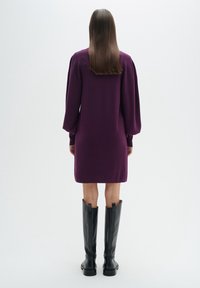Lila långärmad klänning med puffade axlar, knälång, kombinerad med knähöga svarta läderstövlar. Slät textur, minimalistisk design.