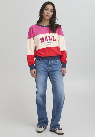 Daudzkrāsains džemperis rozā, krēmu un sarkanā krāsā ar izteiksmīgu "BALL" tekstu un ziliem akcentiem, apvienots ar gaismas mazgātām džinsiem un ikdienišķām kurpēm.