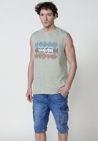 Gaiši zaļš bez piedurknēm krekls ar tekstu "Sērfojot pa viļņiem" un daudzkrāsainu viļņu rakstu. Saskaņots ar līdz ceļiem ziliem cargo šortiem.