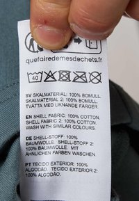 Etikett med skötselråd och materialinformation: 100 % bomull, flerspråkig text, slät yta, rektangulär form, olika tvättsymboler.