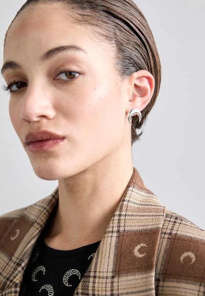 Boucles d'oreilles en forme de croissant argenté sur un mannequin, associées à un blazer marron à carreaux et un haut noir avec des embellissements brillants.