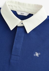 Tamsiai mėlynas polo marškinėliai su baltu medvilniniu apykakle, turintys trikampę atvartą su trimis mygtukais ir mažą baltą siuvinėtą logotipą.