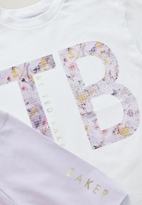 Bijela pamučna majica s velikim ljubičastim cvjetnim sequinskim logotipom "TB". U paru s svijetloplavim kratkim hlačama s zlatnim tekstualnim akcentima.