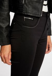 Vrouw in zwarte strakke broek met zichtbare voorrits en cropped zwart leren jasje met zilveren rits.