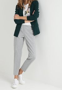 Blazer su misura verde scuro sopra una t-shirt bianca con grafica, abbinato a pantaloni sportivi grigi chiari a sigaretta e sneaker bianche.