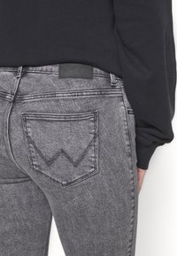 Jeans de mezclilla gris con un acabado texturizado, que cuentan con una costura en forma de V en el bolsillo trasero y una etiqueta de cuero negra en la cinturilla.