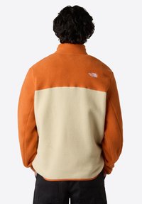 Fleecejack met een oranje bovenkant en een crème onderkant. Heeft een zachte textuur en een hoge kraag. Klein logo op de linkerschouder.