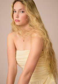 Zonder schouder lichte gele jurk met geplooide stof, een gladde textuur en een off-the-shoulder ontwerp. Lange golvende blonde haren en minimale sieraden.