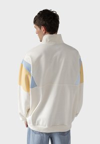 Sudadera blanca de manga larga con diseño de bloques de color en amarillo, azul y blanco. Cuello alto y corte relajado, hecha de una tela suave y texturizada.