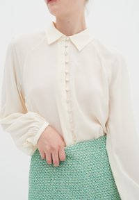 Lys beige bluse med krave, knaplukning foran og samlede ærmer, parret med en tekstureret grøn nederdel med et vævet mønster.