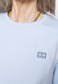 Sudadera azul claro con un cuello redondo, que presenta un pequeño logo rectangular bordado en azul marino oscuro. Textura de algodón suave.