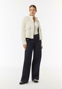 Jasnobeżowa kurtka z wysokim kołnierzem i przednimi kieszeniami, w połączeniu z ciemnoniebieskimi szerokimi dżinsami z ozdobnymi złotymi guzikami. Całość dopełniają czarne buty.