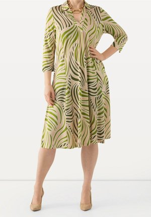 Vrouw die een beige jurk tot op de knie draagt met groene abstracte bladpatronen, driekwart mouwen en beige hoge hakken.