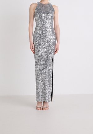 Ærmeløs aftenkjole med sølvpalietter, høj halsudskæring og sideslids, båret med sølvfarvede remsandaler på ensartet baggrund.