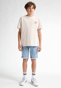 Light beige cotton t-shirt with a small logo, paired with rolled-up light blue denim shorts and white sneakers with black accents.