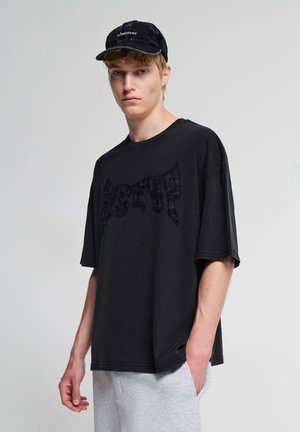 Giovane uomo che indossa un berretto nero "Whatever" e una t-shirt nera oversize con un design sottile e testurizzato, pantaloni della tuta grigi, posato davanti a uno sfondo uniforme.