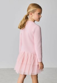 Svetloružové šaty s priliehavým topom a vrstvami priesvitnej tylovej sukne. Mäkká, textúrovaná látka s dlhými rukávmi a zaobleným výstrihom.