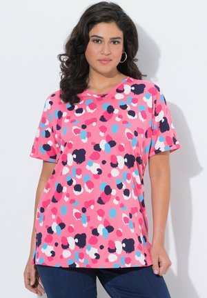 Frau mit dunklem, lockigem Haar, die ein rosa Kurzarmoberteil mit mehrfarbigen abstrakten Flecken und eine dunkelblaue Jeans vor weißem Hintergrund trägt.