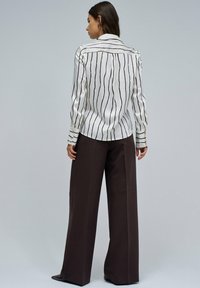 Blusa blanca de manga larga con rayas verticales negras y cuello, combinada con pantalones de pierna ancha color marrón oscuro y accesorios mínimos.