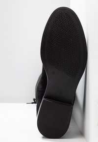 Semelle de chaussure noire avec bande de roulement texturée et indication de taille 4½, photographiée debout sur un fond blanc.