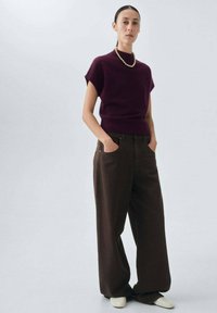 Bordeaux gebreide top met korte mouwen en een opstaande kraag, gecombineerd met losse bruine broeken en witte schoenen. Eenvoudige parelketting als accent.