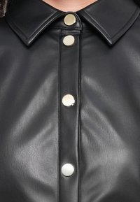 Vêtement en cuir noir avec un col pointu et quatre boutons-pression ronds argentés brillants au centre devant.
