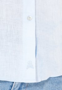 Chemise en lin bleu clair avec fermeture à boutons et détail brodé de crâne et os croisés au bas. Texture lisse, coupe décontractée.